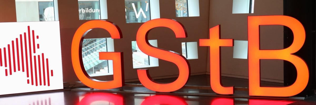 "GStB" in leuchtenden, menschengroßen Buchstaben vor einer Fensterwand.