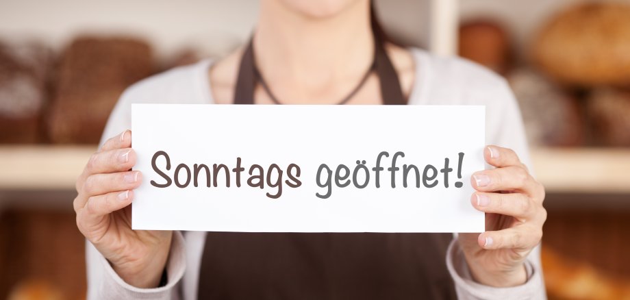 Eine Frau mit Schürze hält ein Schild in den Händen mit der Aufschrift "Sonntags geöffnet!". Sie steht vor einem Regal mit Broten.