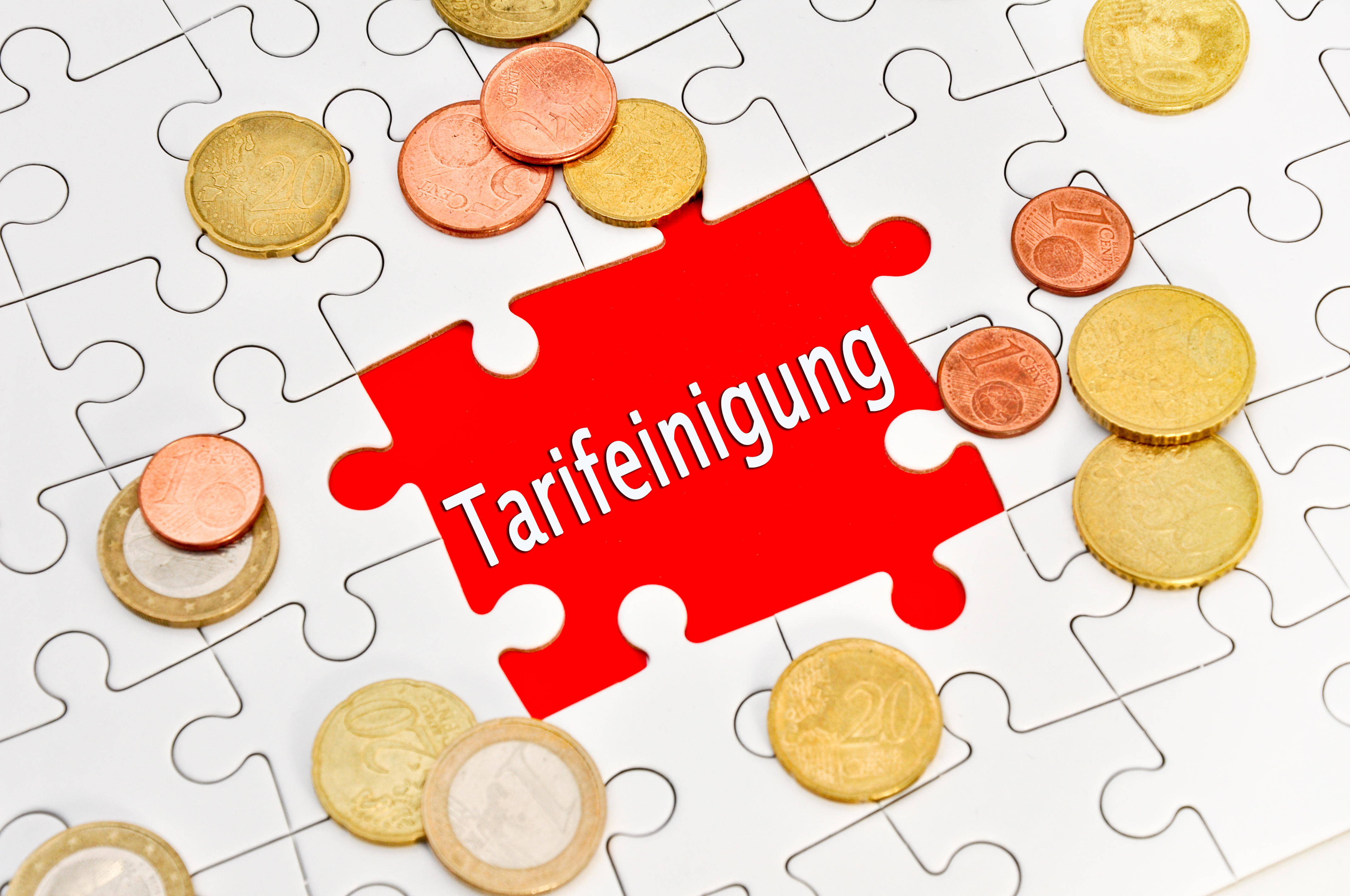 Ein Puzzle, in dessen Mitte ein Puzzleteil fehlt. Dort steht rot hinterlegt  das Wort "Tarifeinigung". Über das Puzzle sind Euro-Cent-Münzen verteilt.