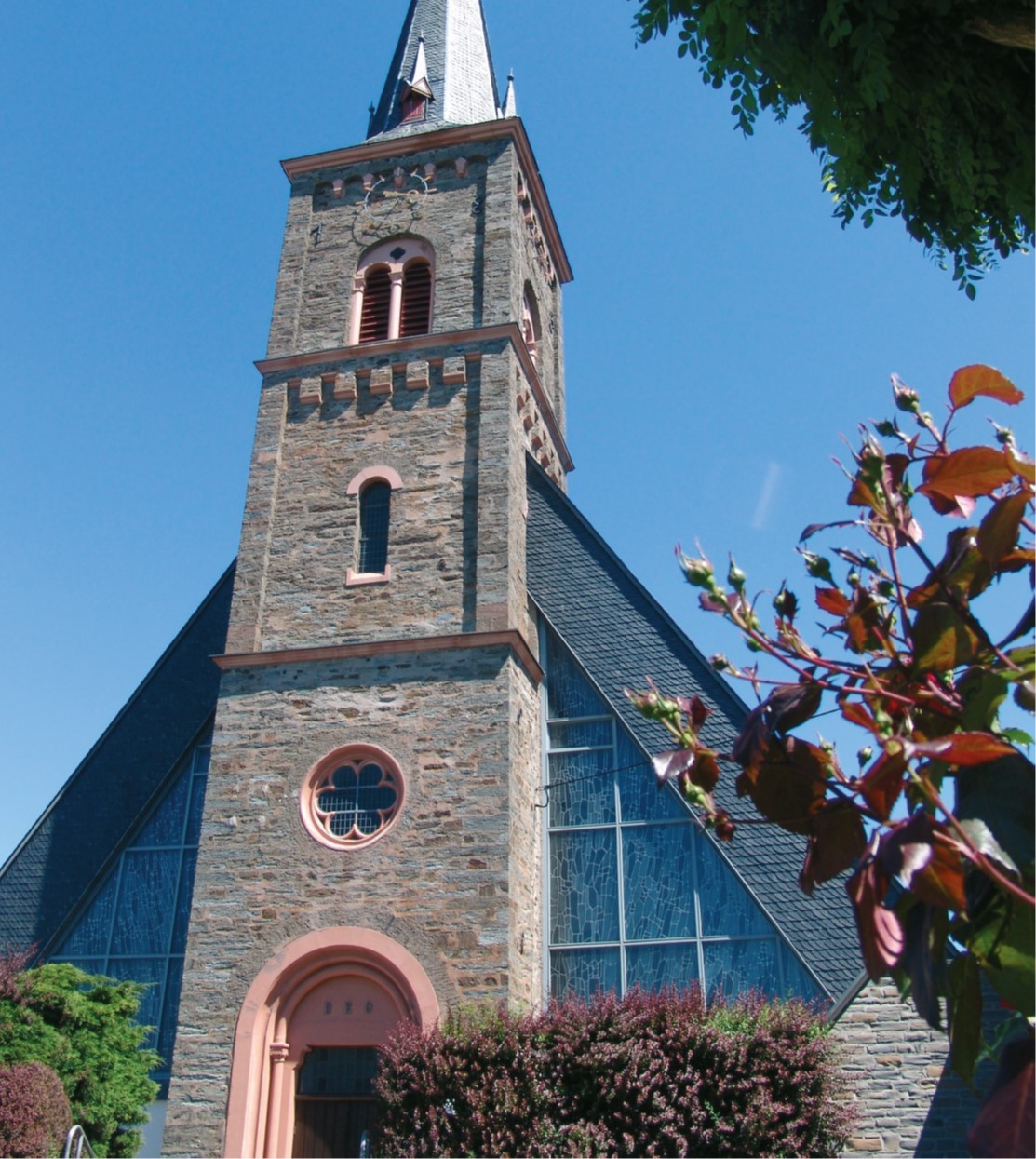 Portal und Kirchturm der katholischen Pfarrkirche St. Andreas der Ortsgemeinde Longkamp vor blauem Himmel.