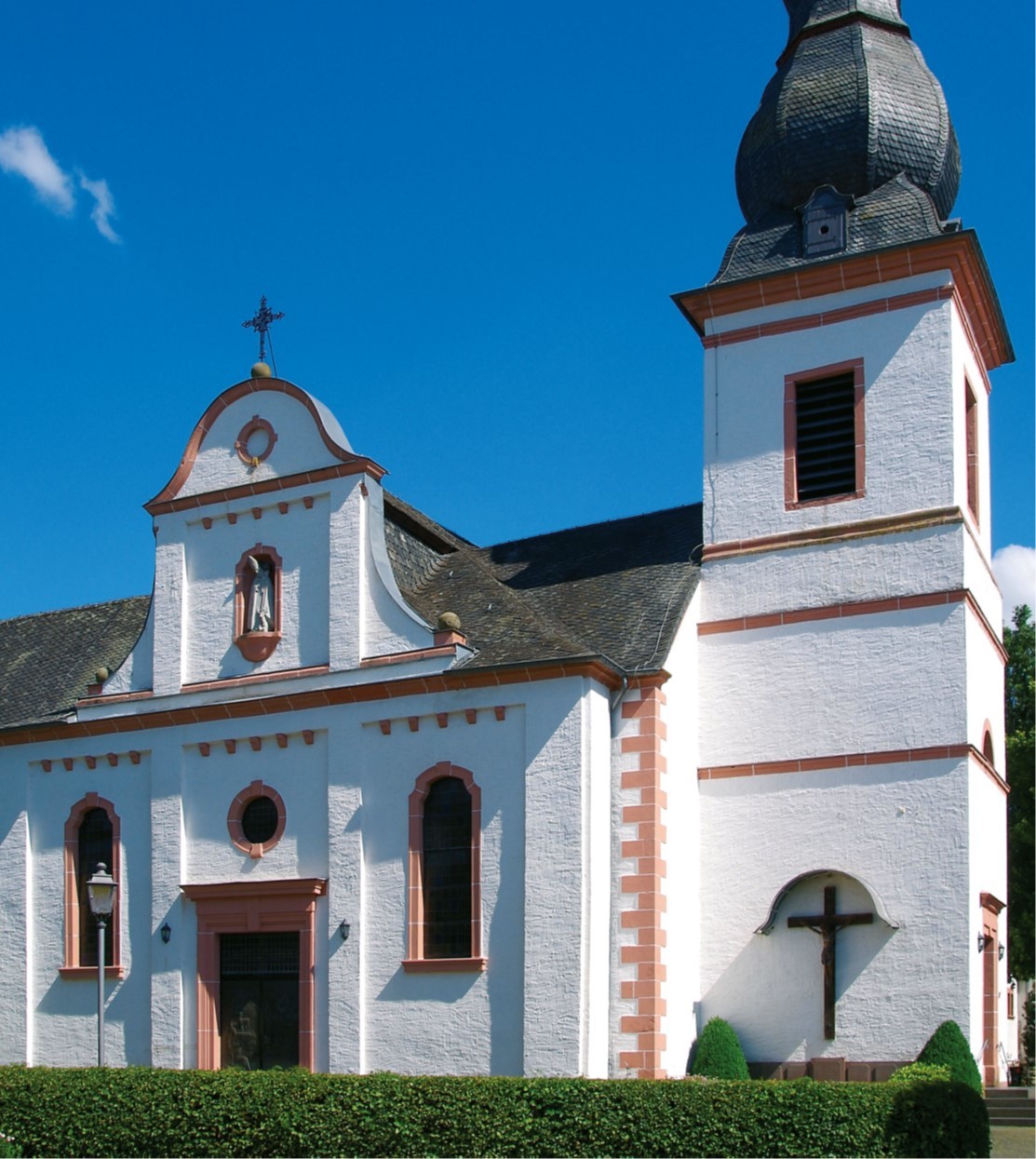  Eingang und Kirchturm der Pfarrkirche Sankt Hubertus vor blauem Himmel.