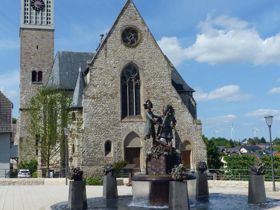 Lebendiges Postkarten-Motiv: Der Drei-Grazien-Brunnen markiert die Mitte von Zornheim und verkörpert das Faible der Einwohner für sinnliche Genüsse – der Wein genießt einen hohen Stellenwert. Erhaben steht die Katholische Kirche im Hintergrund. Rechte: Alexander Sell, Nieder-Olm