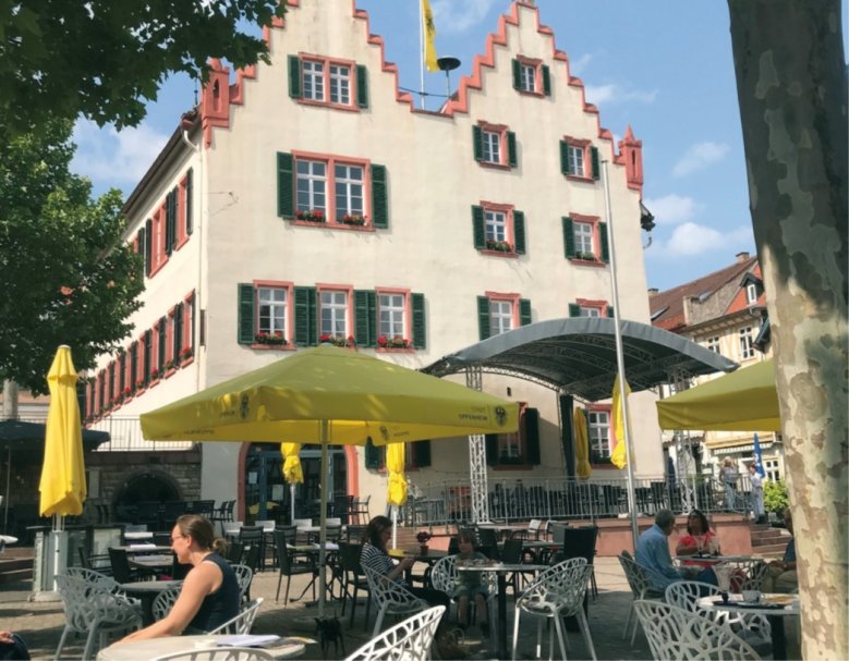 Unter der Innenstadt befindet sich das „Oppenheimer Kellerlabyrinth“ mit 40 Kilometern Länge und zahlreichen Kellern und Gängen. Der Marktplatz von Oppenheim mit dem Rathaus im Hintergrund. Vor dem Rathaus befindet sich eine Bühne. Im Vordergrund sitzen Menschen im Freien in einem Café an runden Tischen und unterhalten sich.