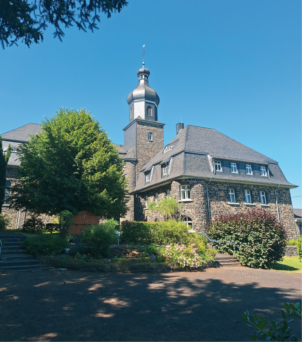 Herz-Jesu-Kirche aus Basaltstein in der Ortsgemeinde Langenhahn umgeben von Bäumen und vor blauem Himmel.