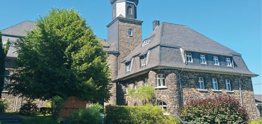 Herz-Jesu-Kirche aus Basaltstein in der Ortsgemeinde Langenhahn umgeben von Bäumen und vor blauem Himmel.