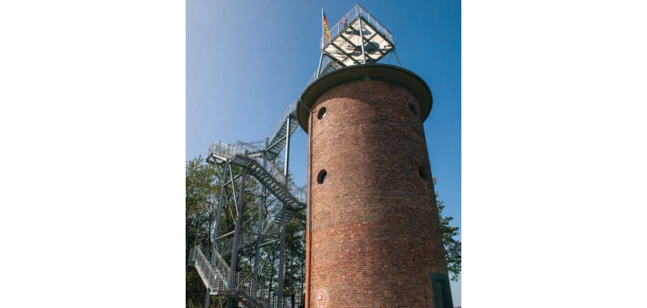 Wasserturm mit Backsteinfassade, Aussichtsplattform und seitlicher Metalltreppe vor blauem Himmel und mit Deutschlandflagge auf der Plattform.