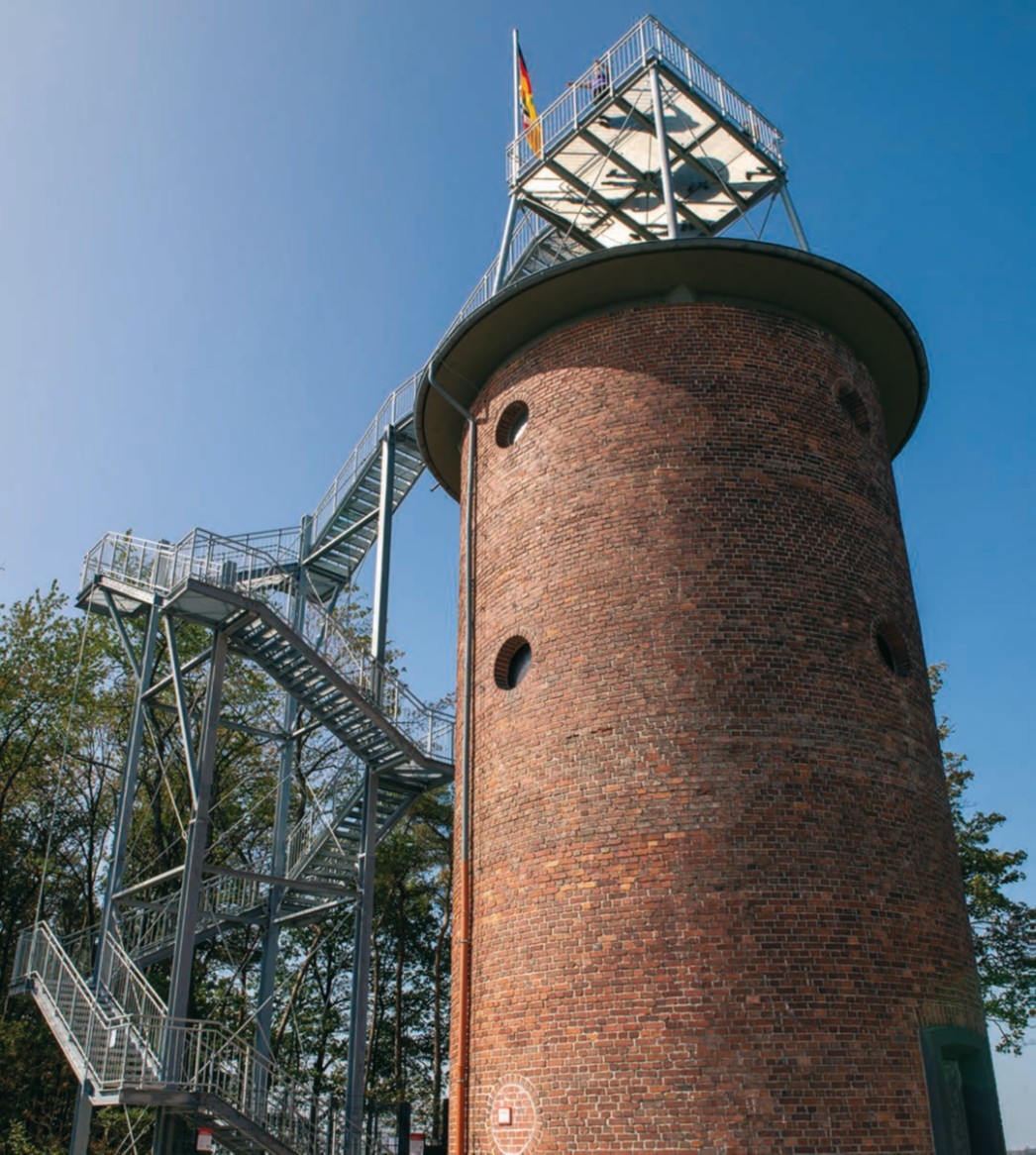 Wasserturm mit Backsteinfassade, Aussichtsplattform und seitlicher Metalltreppe vor blauem Himmel und mit Deutschlandflagge auf der Plattform.