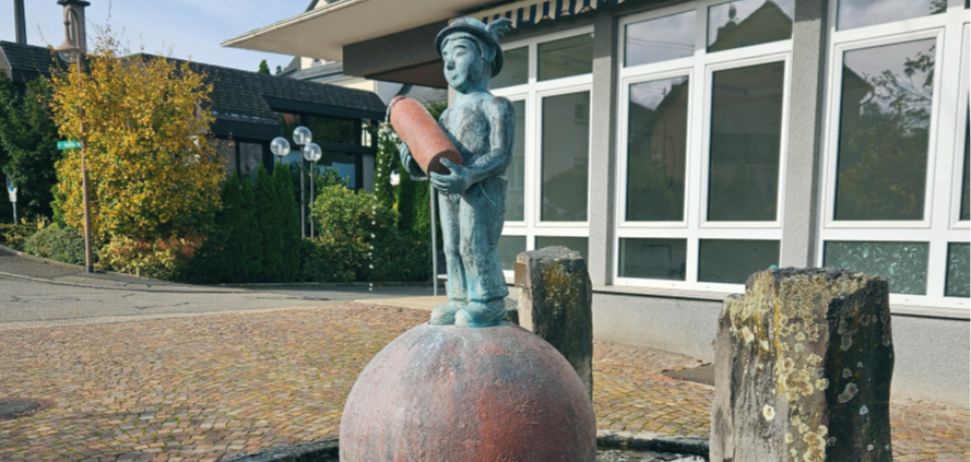 Brunnen auf einem gepflasterten Platz in der Ortsgemeinde Mogendorf. In der Mitte des Brunnens befindet sich eine große Kugel auf der eine Männchen mit einem Gefäß in der Hand steht, aus dem Wasser in den Brunnen fließt. 