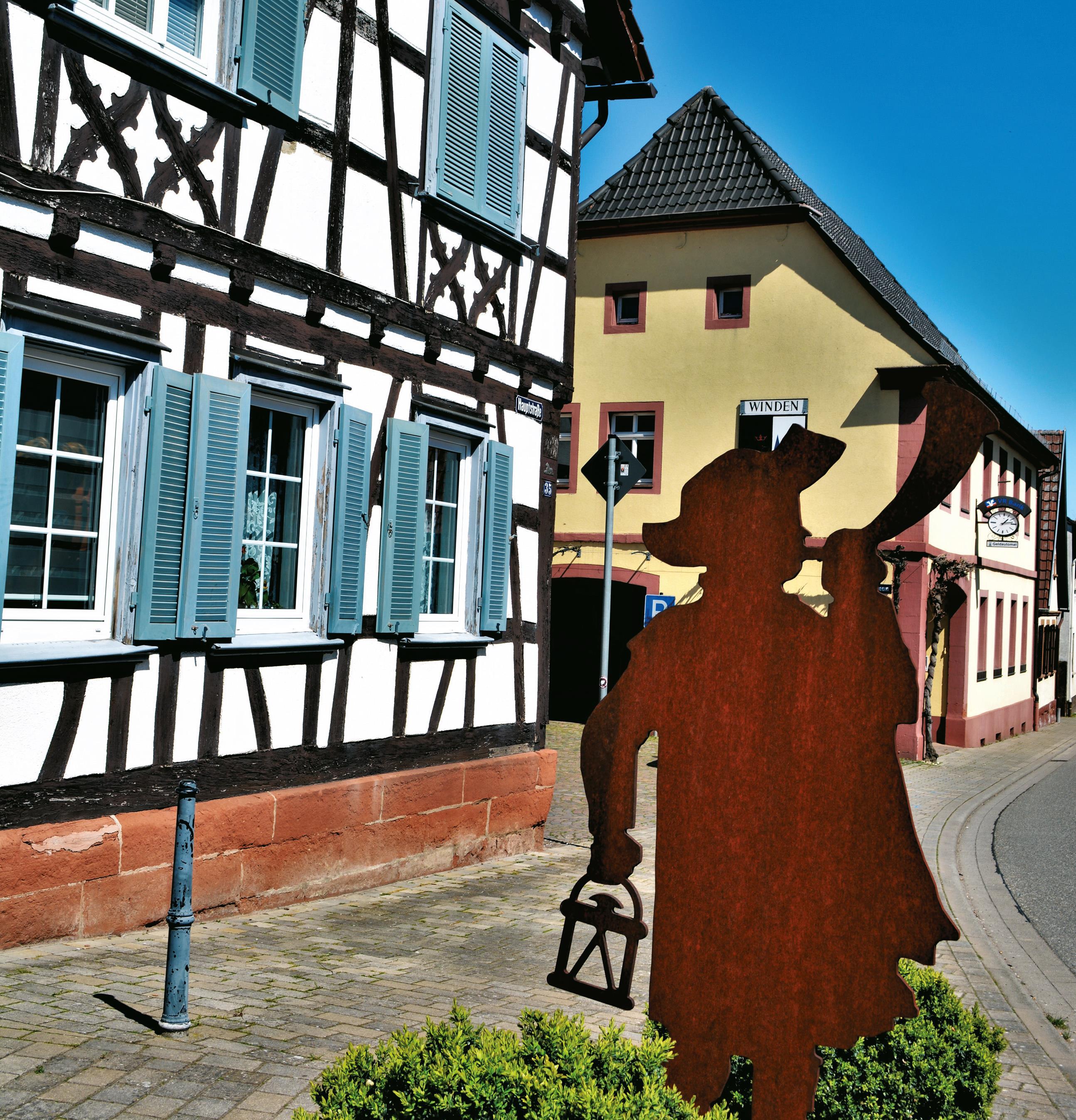 Ortskern der Gemeinde Winden mit alten Fachwerkhäusern. Im Vordergrund ist die Metallskulptur eines Nachtwächters zu sehen.