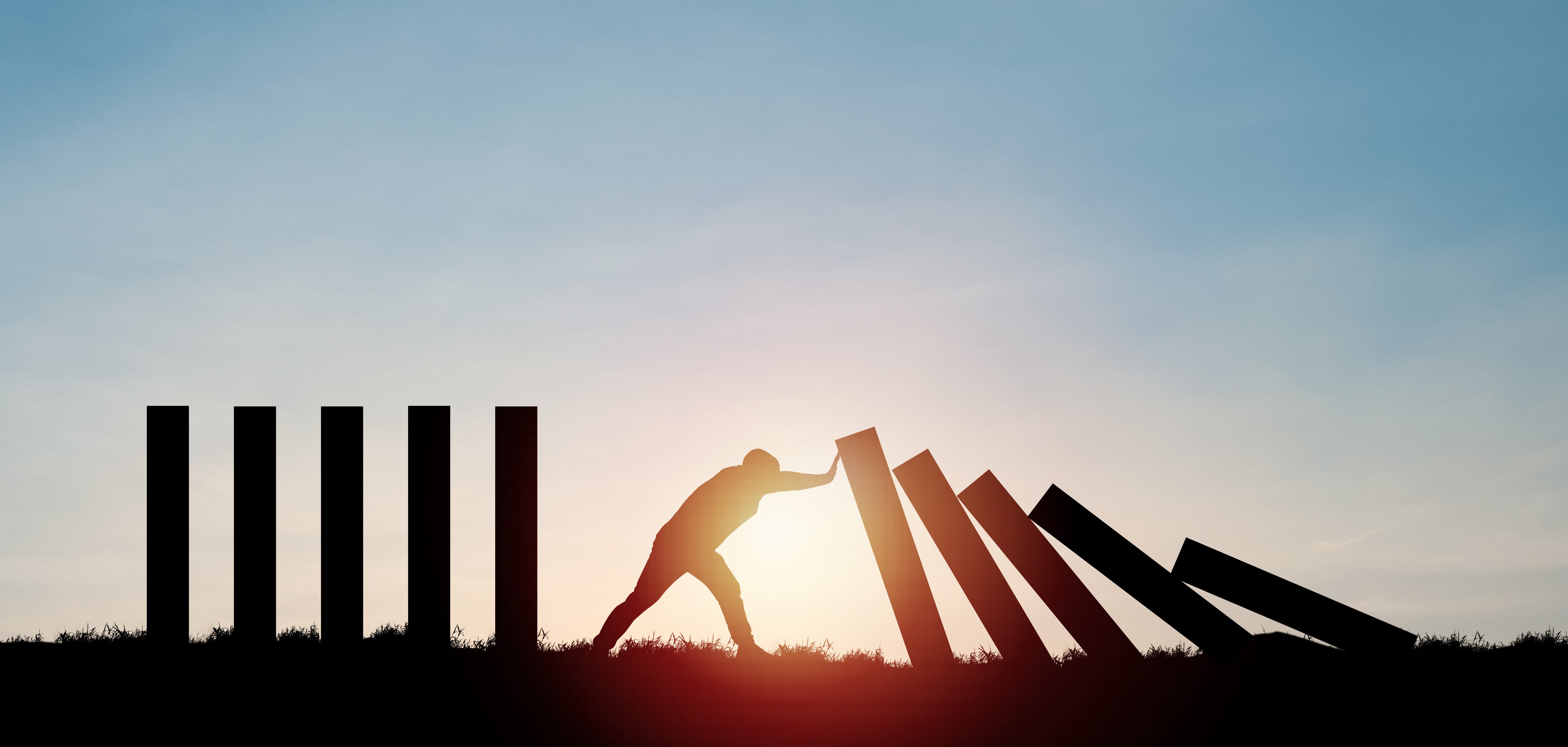 Das Bild zeigt die Silhouette eines Mannes vor einem blauen Himmel. Er versucht, große fallende Dominosteine aufzuhalten.