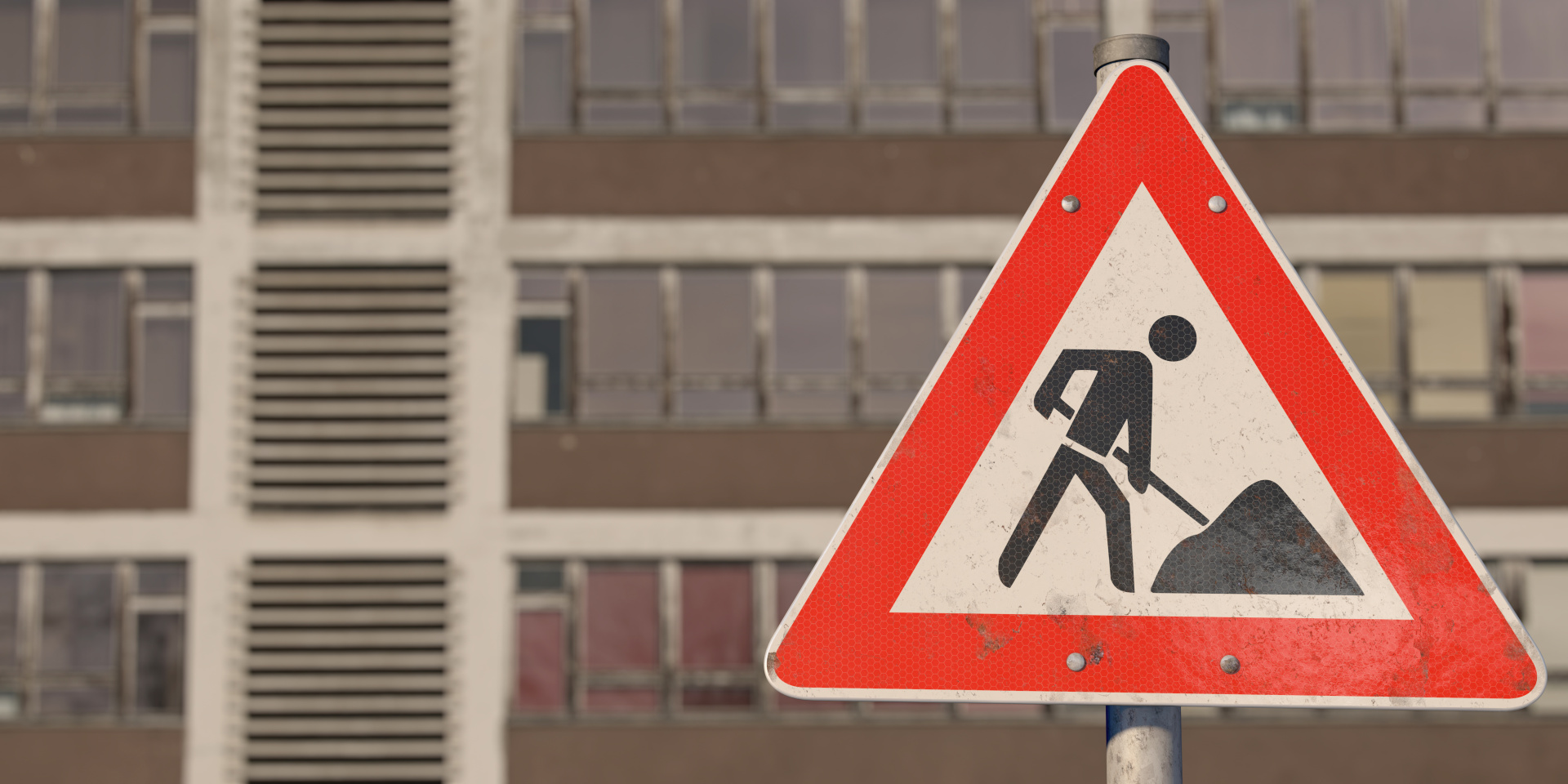 Marodes Schulgebäude. Davor ein dreieckiges Warnschild mit rotem Rand. In der Mitte ist eine schwarze Figur abgebildet, die mit einer Schaufel arbeitet. Bedeutung: Achtung, Baustelle.