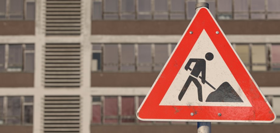 Marodes Schulgebäude. Davor ein dreieckiges Warnschild mit rotem Rand. In der Mitte ist eine schwarze Figur abgebildet, die mit einer Schaufel arbeitet. Bedeutung: Achtung, Baustelle.