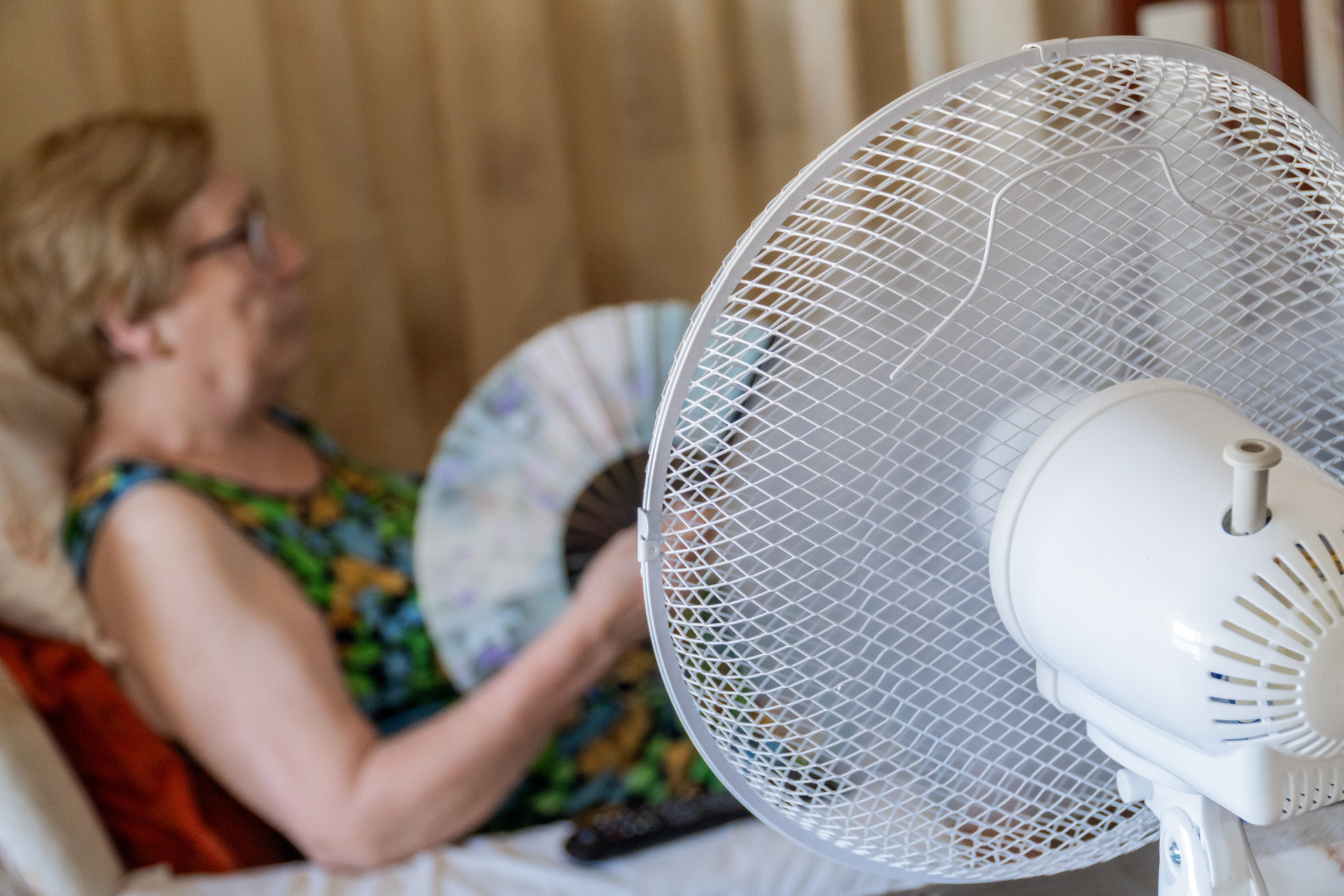 Ein elektrischer Ventilator kühlt eine ältere Frau, die im Wohnzimmer auf dem Sofa sitzt.