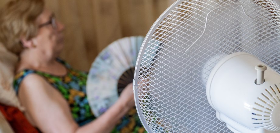 Ein elektrischer Ventilator kühlt eine ältere Frau, die im Wohnzimmer auf dem Sofa sitzt.