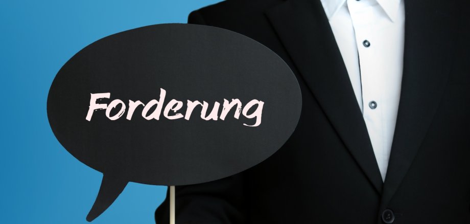 Mann im Anzug hält Sprechblase in der Hand. Der Text Forderung steht im Schild. Blauer Hintergrund.