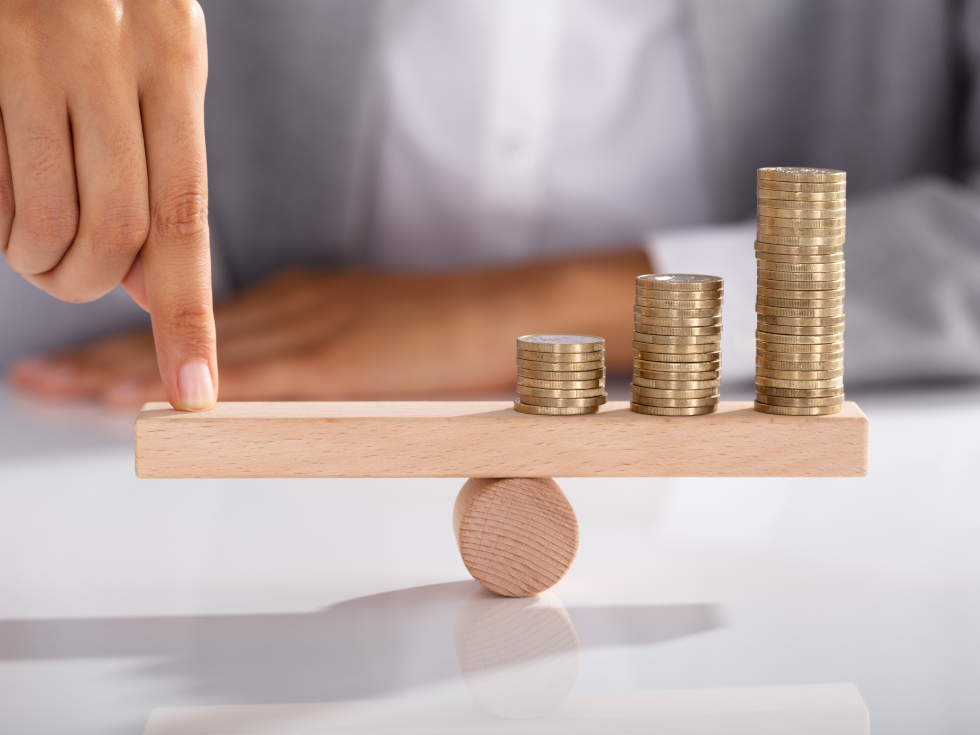 Ein Finger drückt auf eine Holzstange, die auf einer runden Rolle liegt. Auf der anderen Seite der Stange liegen Münzstapel. Die Münzen sind unterschiedlich hoch. Das Bild zeigt ein Gleichgewicht zwischen dem Finger und dem Geld.