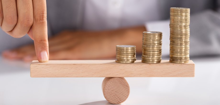 Ein Finger drückt auf eine Holzstange, die auf einer runden Rolle liegt. Auf der anderen Seite der Stange liegen Münzstapel. Die Münzen sind unterschiedlich hoch. Das Bild zeigt ein Gleichgewicht zwischen dem Finger und dem Geld.