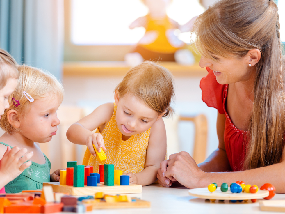 Kitakinder sitzen mit Erzieherin an einem Tisch und spielen mit Holzspielzeug.