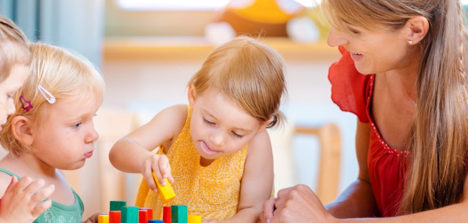 Kitakinder sitzen mit Erzieherin an einem Tisch und spielen mit Holzspielzeug.