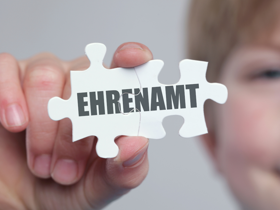 Ein Kind hält ein Puzzleteil in den Vordergrund mit der Aufschrift "Ehrenamt". 