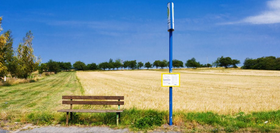 Eine leere Holzbank und ein Haltestellenschild vor einem Feld und blauem Himmel