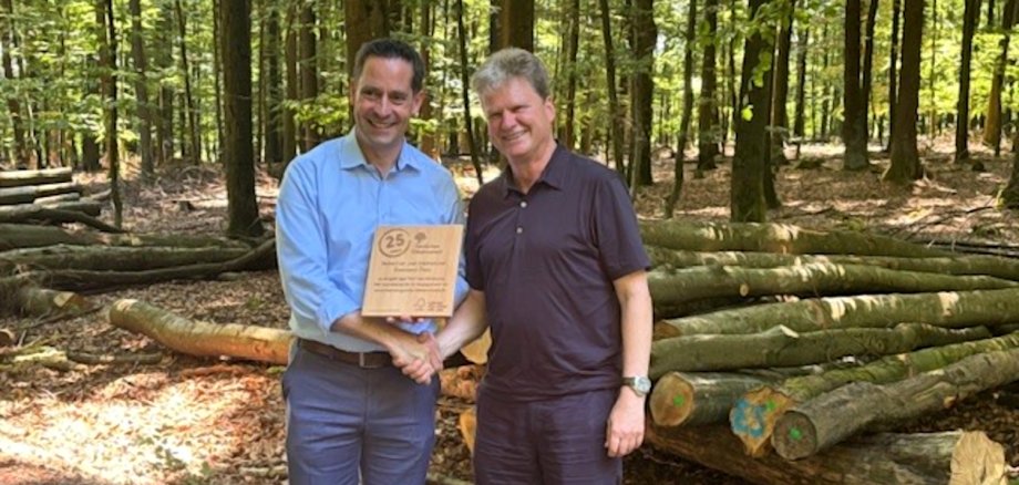 Moritz Petry, Geschäftsführendes Vorstandsmitglied des GStB, und Alfred Schumm, Geschäftsführer von FSC Deutschland, mit Urkunde vor einem Holzstapel im Wald.