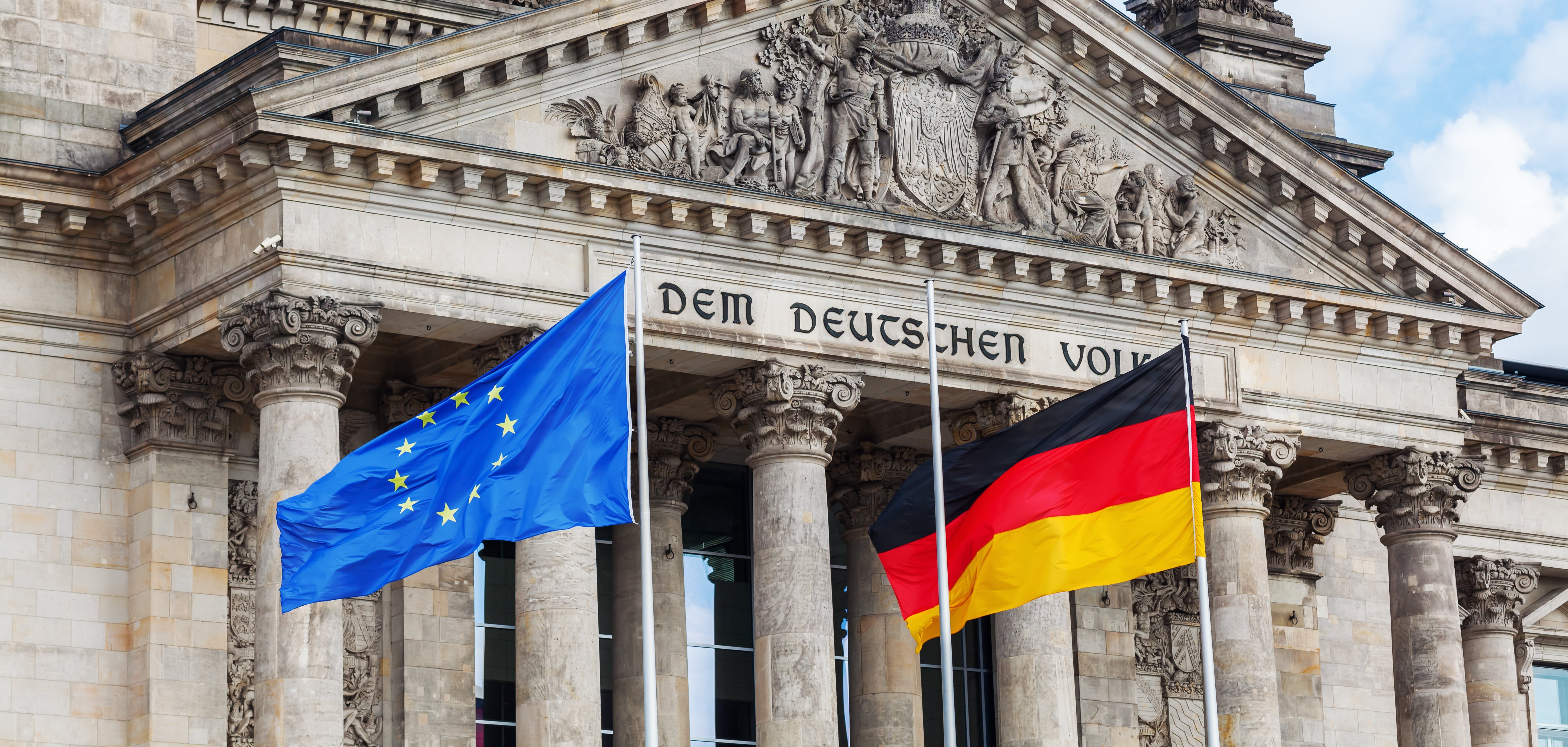 Der Deutsche Bundestag in Berlin. Davor wehen die Europa- und die Deutschlandflagge im Wind. 