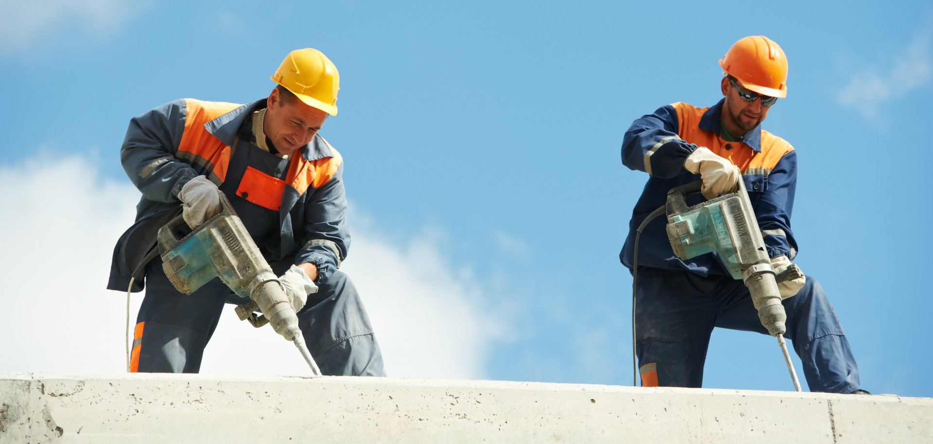 Zwei Bauarbeiter mit Schlagbohrmaschine auf der Baustelle vor blauem Himmel.