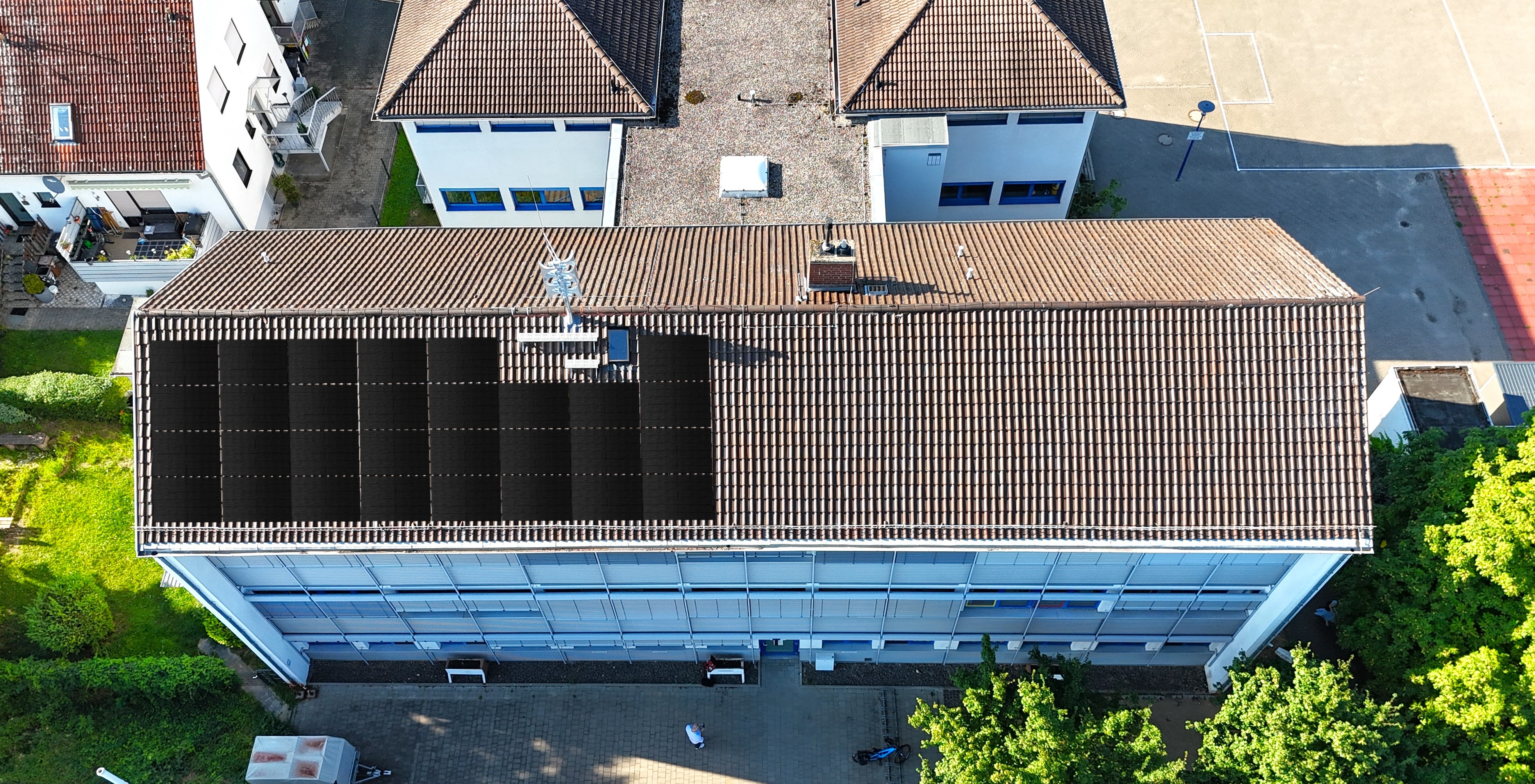 Auf dem Dach der Grundschule Gau-Bischofsheim ist geplant, neben der eigenen Photovoltaikanlage eine weitere zur Volleinspeisung einer Energiegenossenschaft zu errichten, der die Verbandsgemeinde beigetreten ist. Zudem ein bis zwei Windräder. Die Luftaufnahme zeigt das Dach der Grundschule Gau-Bischofsheim, auf dem eine Photovoltaik-Anlage simuliert ist.