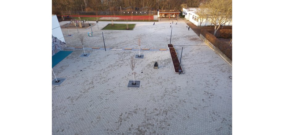 Gepflasterter Schulhof mit neu gepflanzten Bäumen, Grünflächen sowie einem Spiel- und einem Sportplatz.